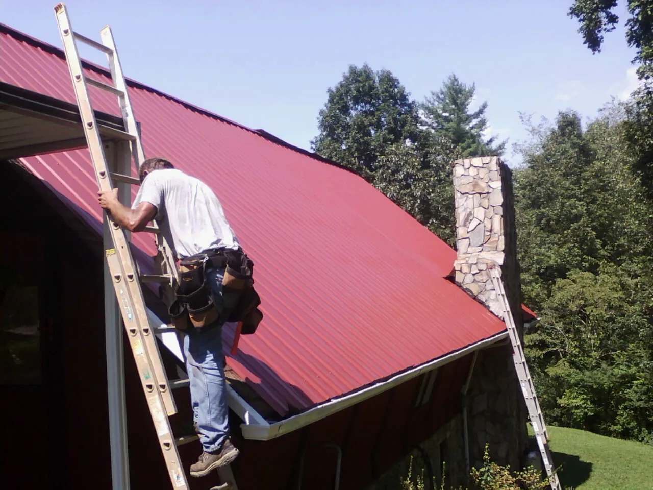 Roofer on a ladder beside a red metal roof and stone chimney — Soffit & Fascia Repair by Roofing Brothers in Alma